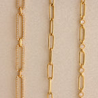 Three gold chain necklaces—a Pave Link Chain Necklace with sparkling diamonds, a plain chain, and one adorned with small round gems—are displayed on a neutral background.