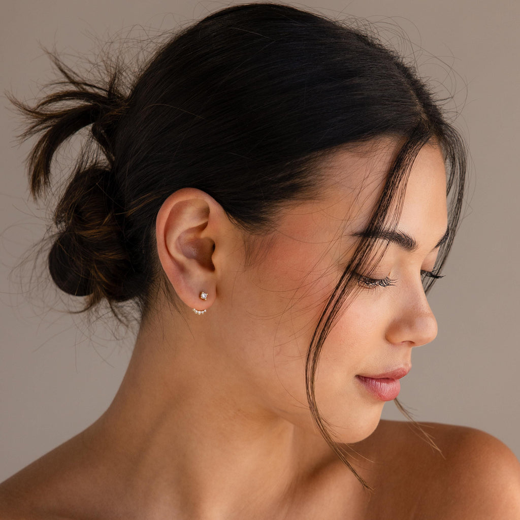 A woman with dark hair in a messy bun looks down, wearing Round Diamond Ear Jackets in 18K Gold and natural makeup.