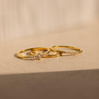 The Trio Diamond Stacking Ring Set—three delicate gold rings with sparkling diamonds—rests on a beige surface, glittering beautifully in warm natural light.