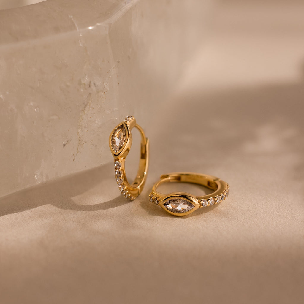 A pair of gold Marquise Pave Huggies adorned with bezeled marquise gemstones and sparkling diamonds rest on a light beige surface near a crystal—ideal for lovers of elegant, art deco-inspired earrings.