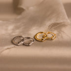 Two pairs of Marquise Pave Huggies hoop earrings—one in silver and one in gold with art deco texture—displayed on beige satin fabric.