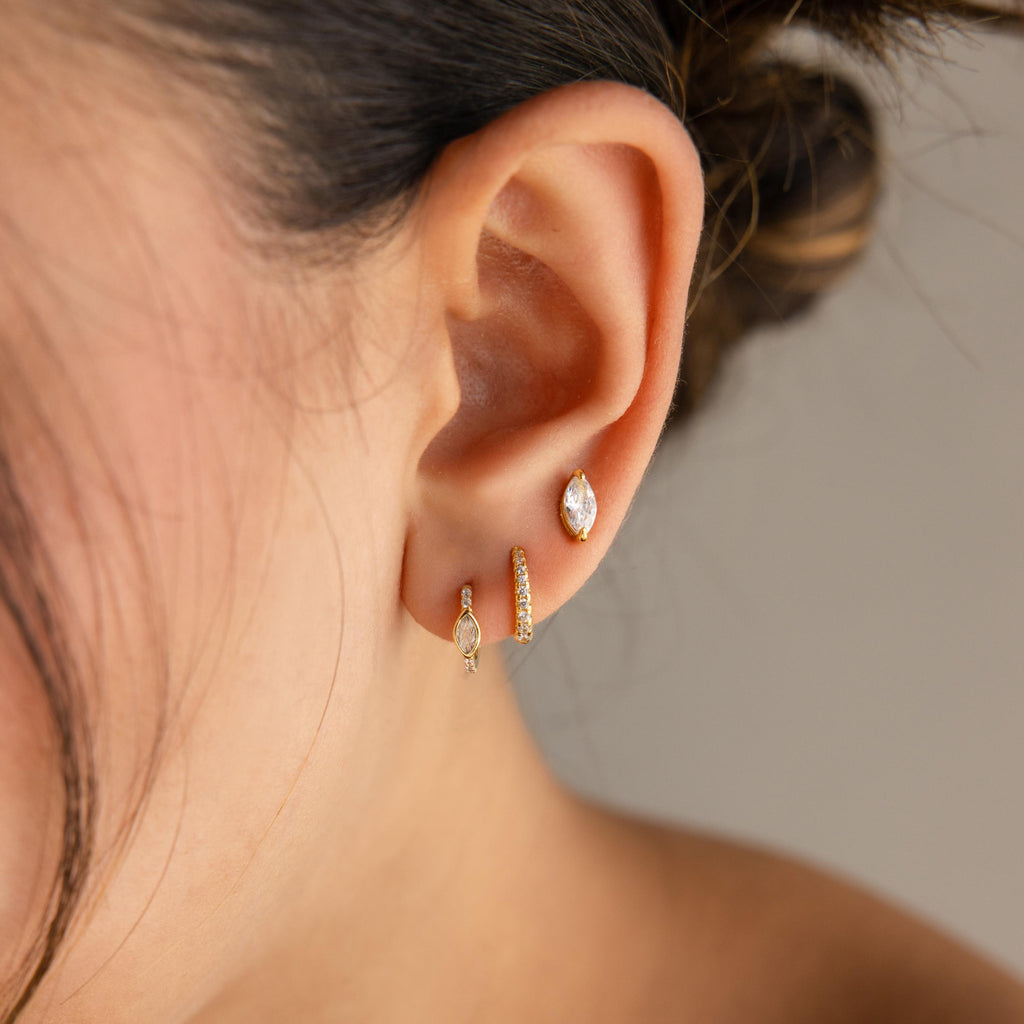 A close-up of an ear wearing three gold earrings: a hoop, a simple stud, and the Marquise Pave Huggies inspired by art deco design.