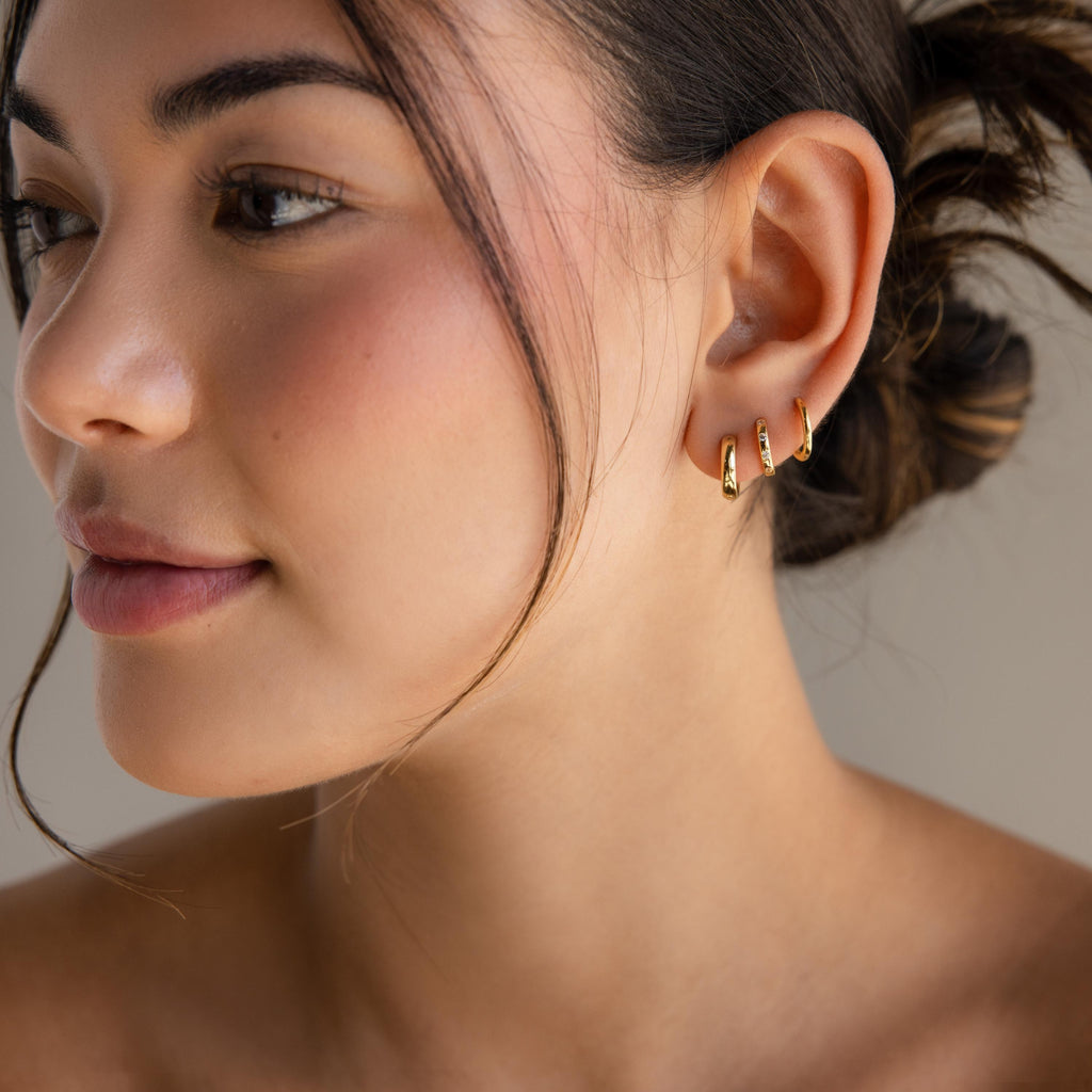 Woman with soft makeup and an updo wears three small gold huggie hoops, including a pair of Mini Diamond Sprinkle Huggies, on her left ear.