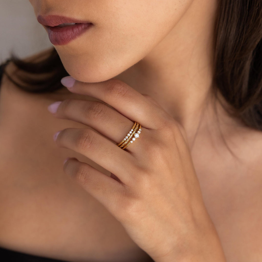 A woman with natural makeup and nude lips touches her face, showcasing the Trio Diamond Stacking Ring Set adorned with small stones on her finger.
