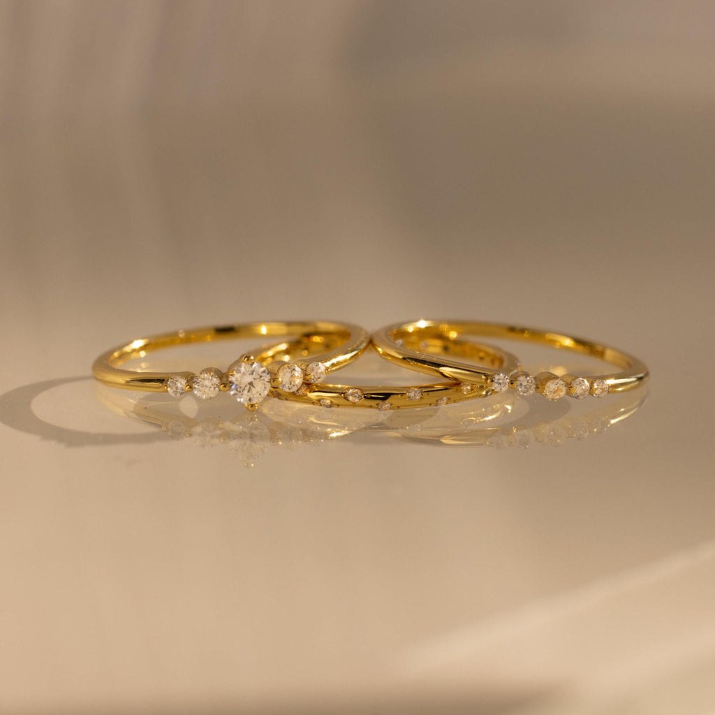 The Trio Diamond Stacking Ring Set, featuring delicate gold bands accented with small diamonds, rests on a smooth, reflective surface bathed in warm light.