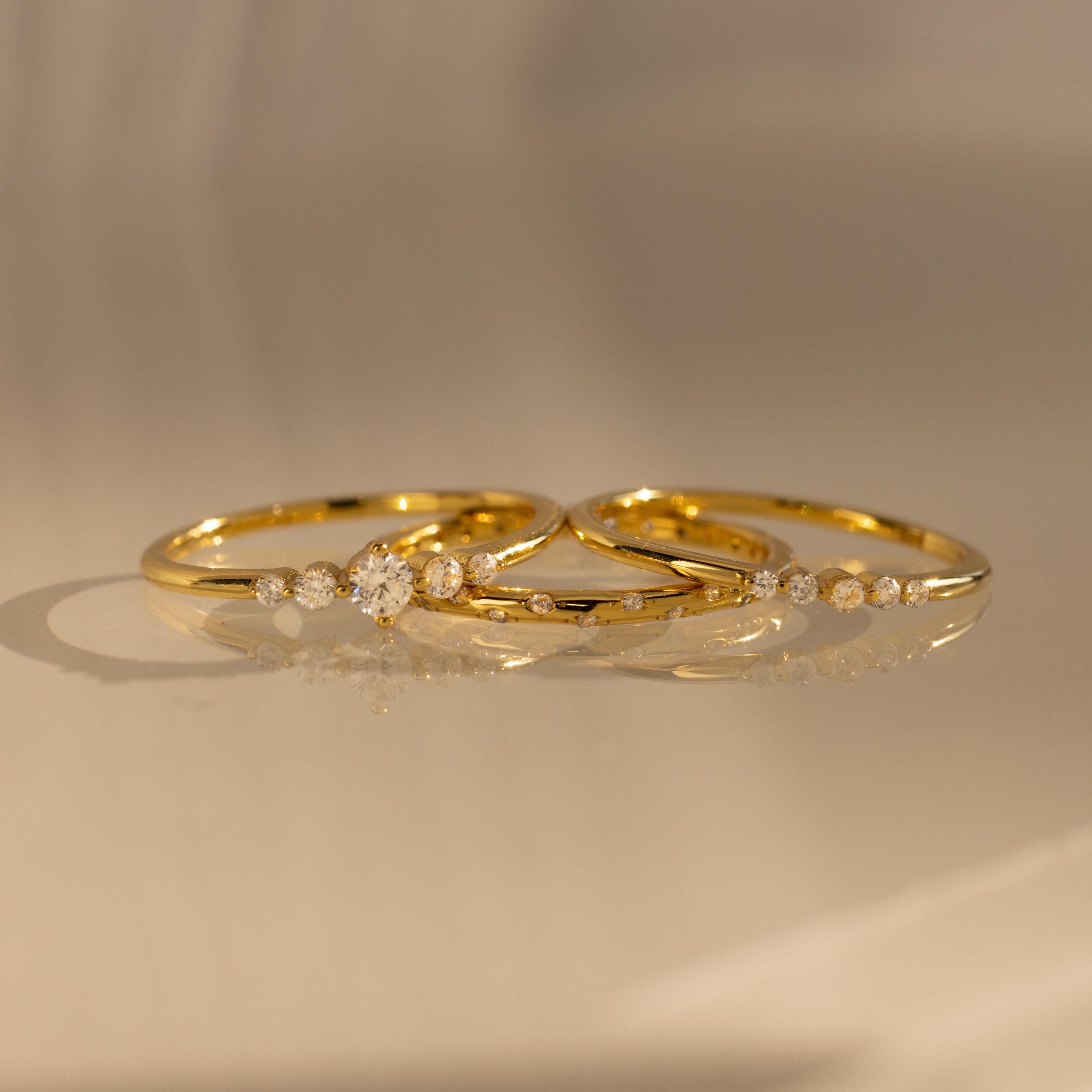The Trio Diamond Stacking Ring Set, featuring delicate gold bands accented with small diamonds, rests on a smooth, reflective surface bathed in warm light.