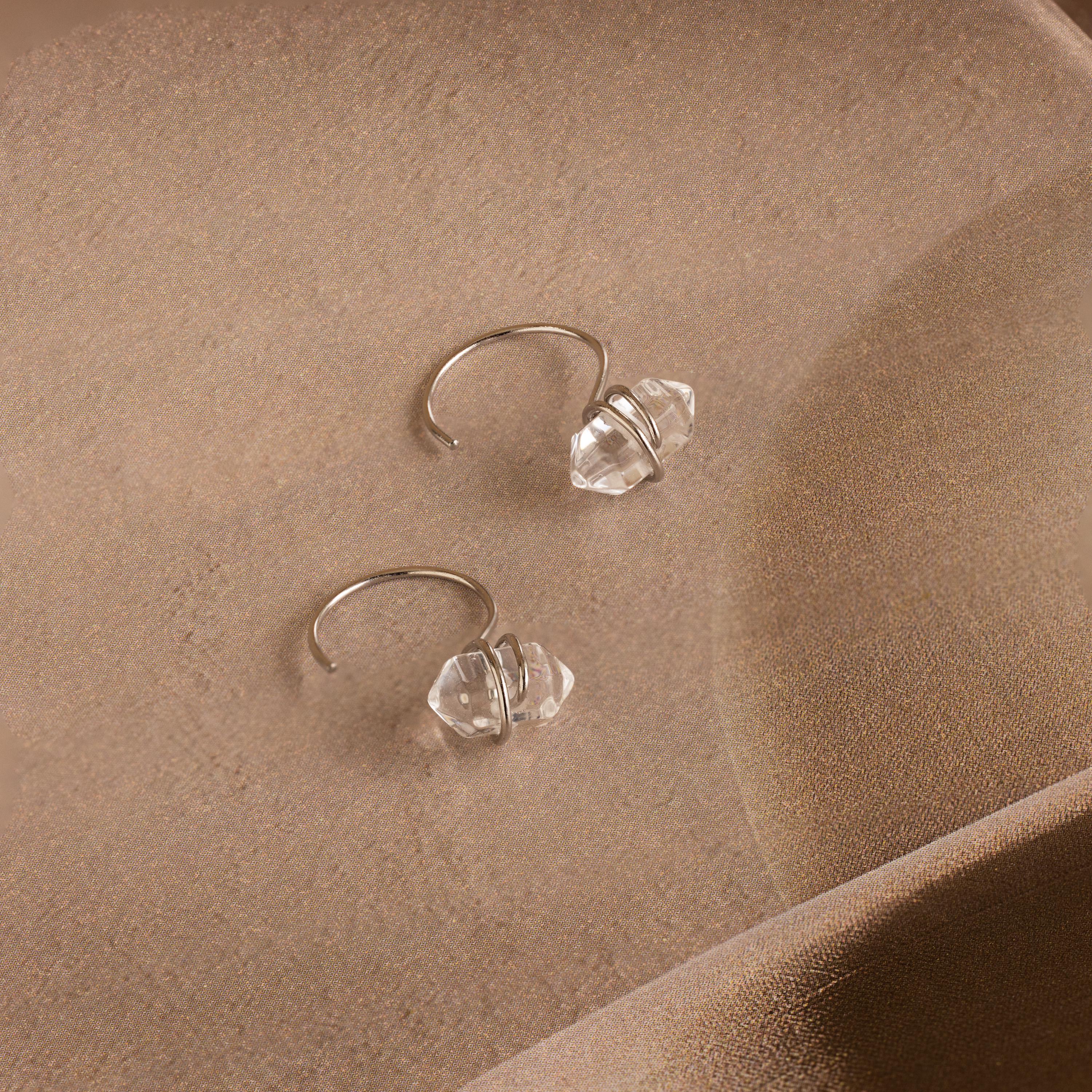The Herkimer Diamond Threaders, nature-inspired silver hoop earrings with clear crystal beads, are displayed on a soft beige fabric background.