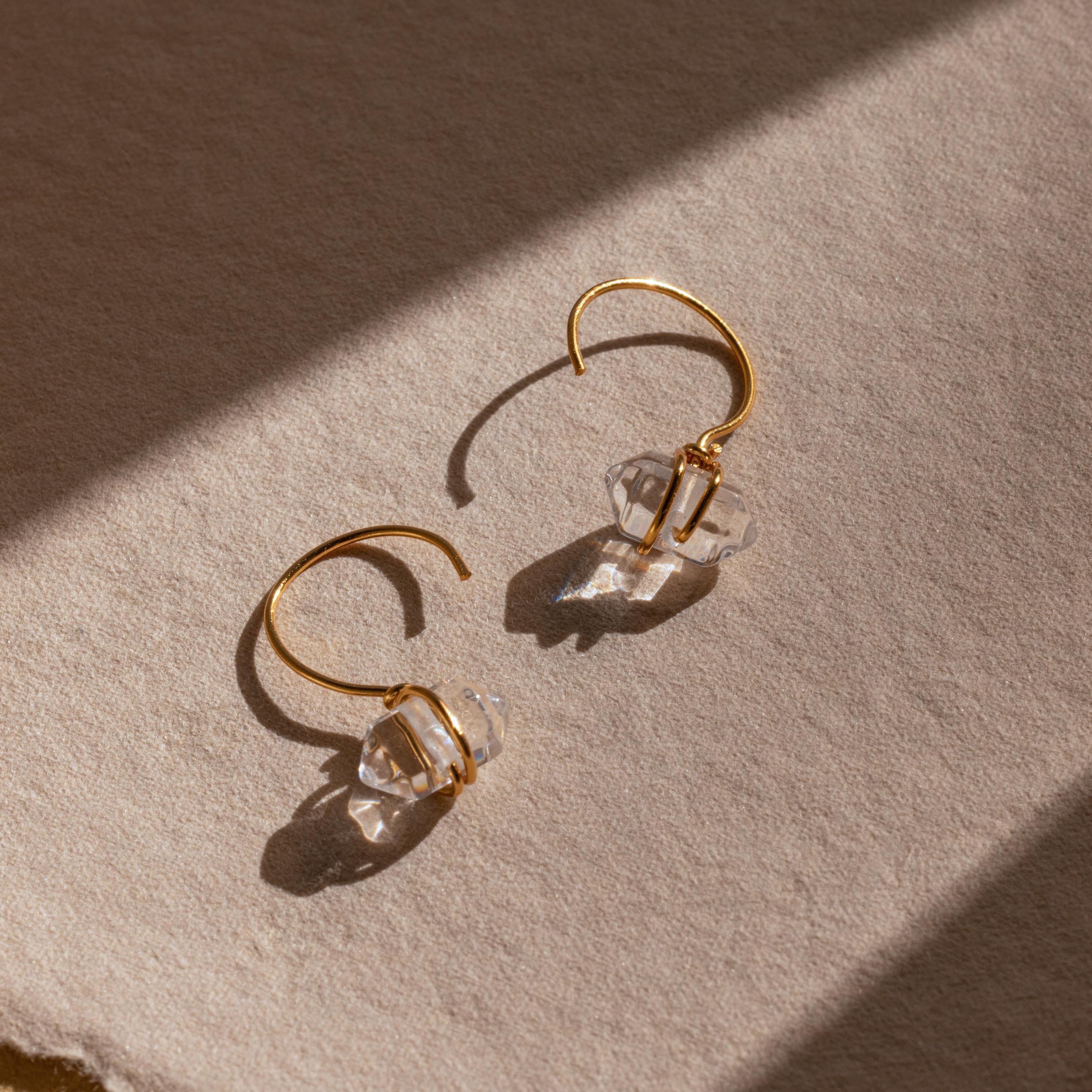 Herkimer Diamond Threaders, featuring clear crystal accents and a nature-inspired design, rest on textured beige fabric bathed in sunlight.
