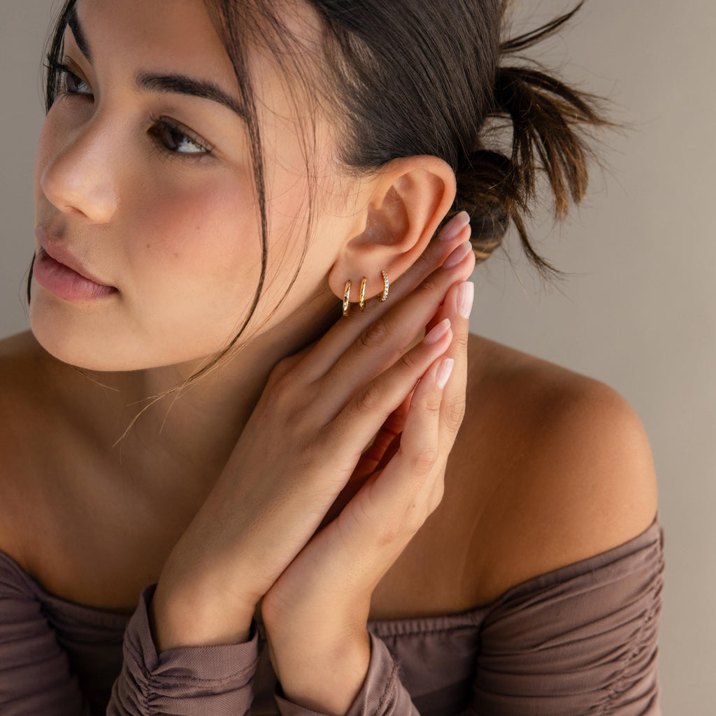 A woman with dark hair in a bun, wearing an off-shoulder brown top, delicately touches her ear adorned with Classic Diamond Sprinkle Huggies.