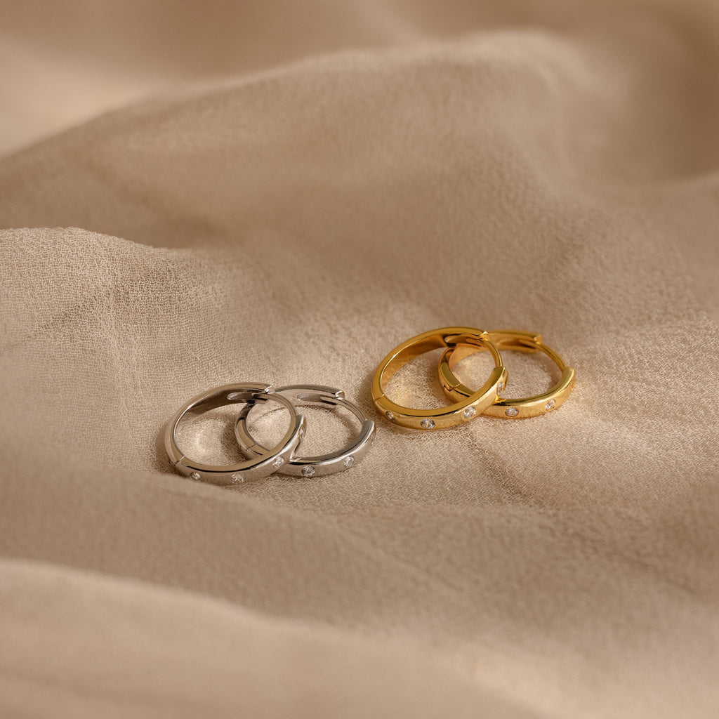 Two pairs of Classic Diamond Sprinkle Huggies, in silver and gold, are displayed on a beige fabric background.
