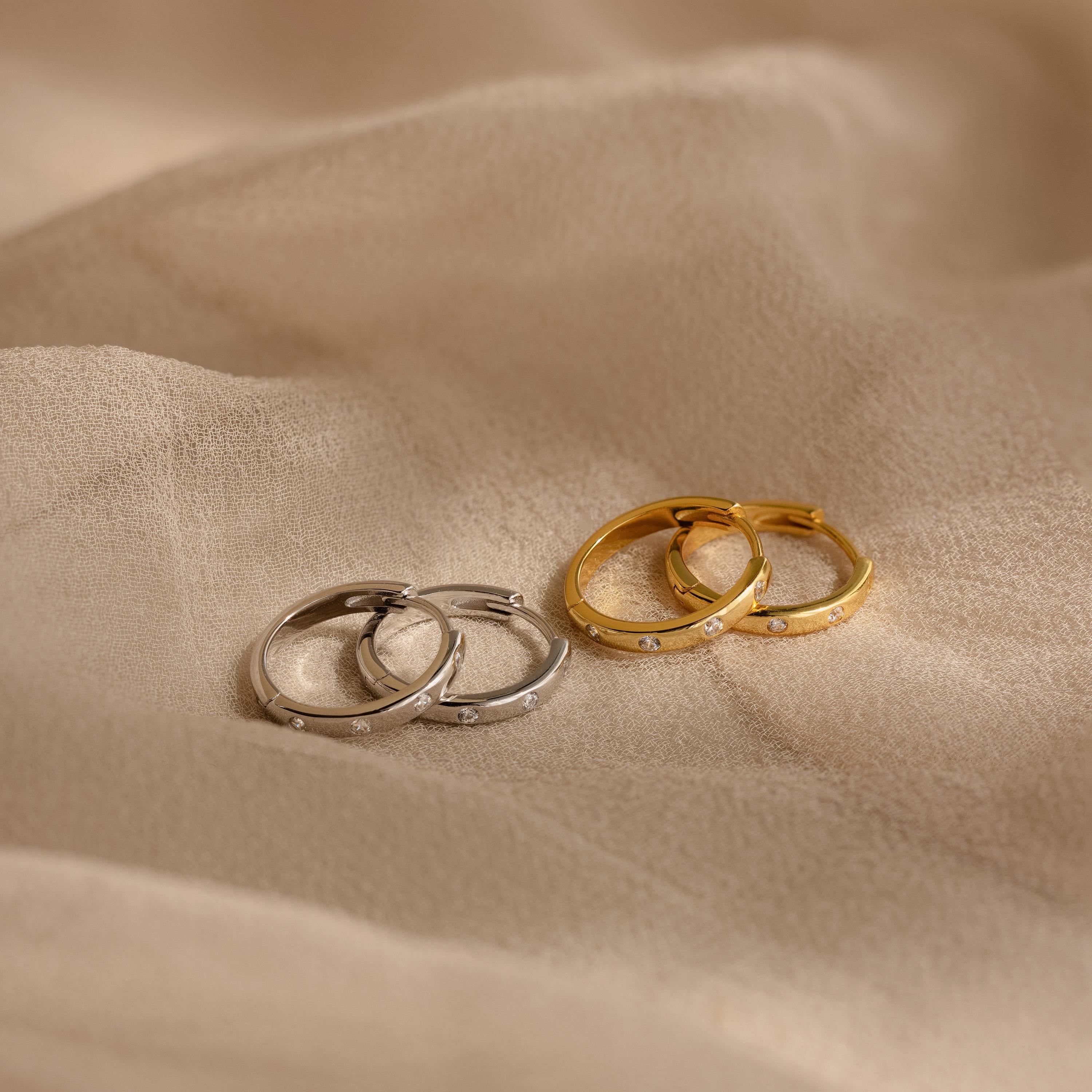 Two pairs of Classic Diamond Sprinkle Huggies, in silver and gold, are displayed on a beige fabric background.