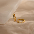 Two gold rings with small diamonds rest on a beige fabric surface near a white marble slab, perfectly complementing your Classic Diamond Sprinkle Huggies for a timeless look.