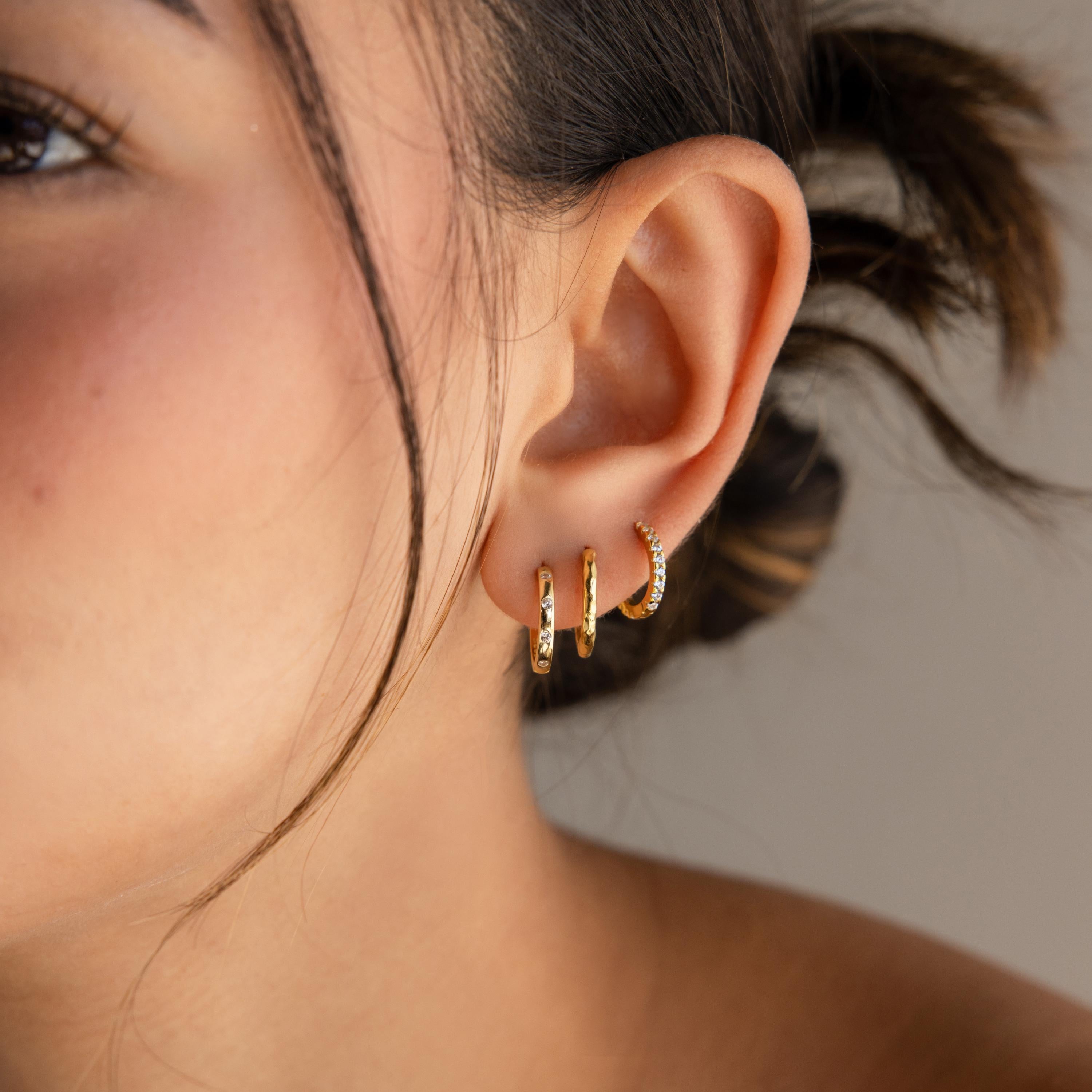 Close-up of a woman's ear wearing three small gold hoop earrings, featuring Classic Diamond Sprinkle Huggies with textured and jeweled art deco designs.