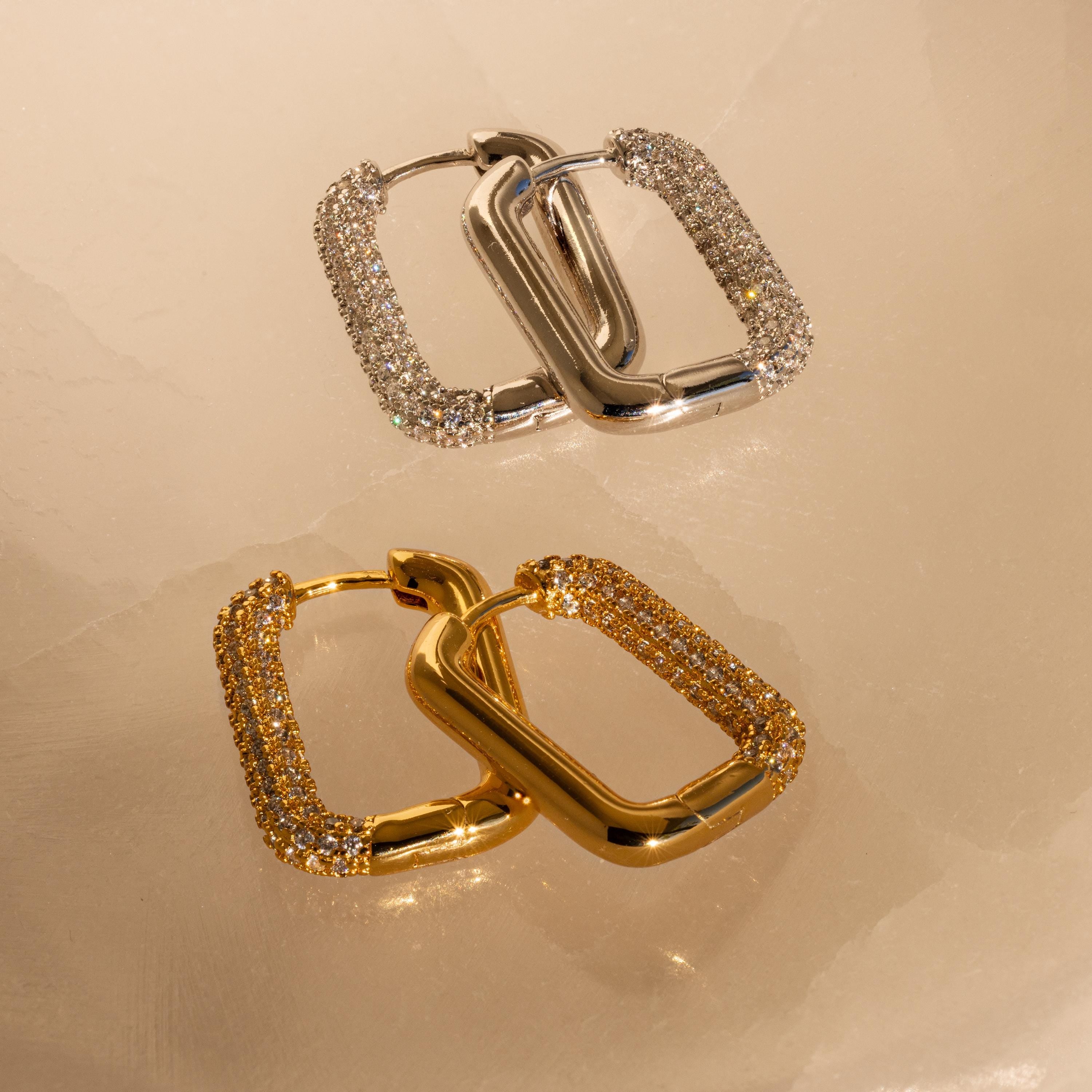 The Chunky Pave Link Earrings, with sparkling stones and a bold gold and silver geometric design, rest on beige marble for a modern take on classic style.