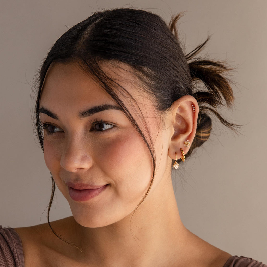 A woman with brown hair in a low bun, smiling softly while wearing Chandelier Diamond Huggies and a taupe top.