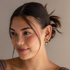 A woman with brown hair in a low bun, smiling softly while wearing Chandelier Diamond Huggies and a taupe top.