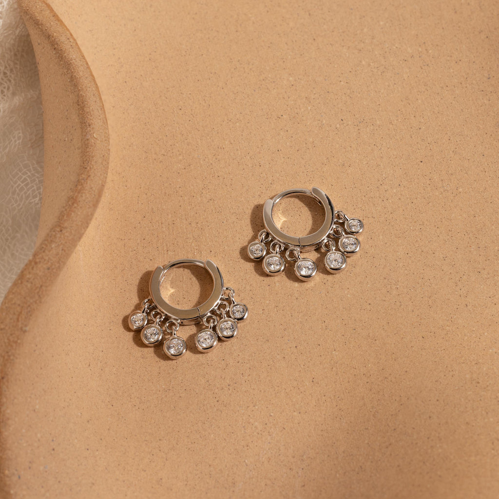 The Chandelier Diamond Huggies shimmer on a tan surface, showcasing timeless elegance and an art deco-inspired charm.