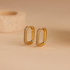Chunky Pave Link Earrings featuring gold geometric hoops with sparkling pavé stones, displayed upright on a beige surface against a blurred background.