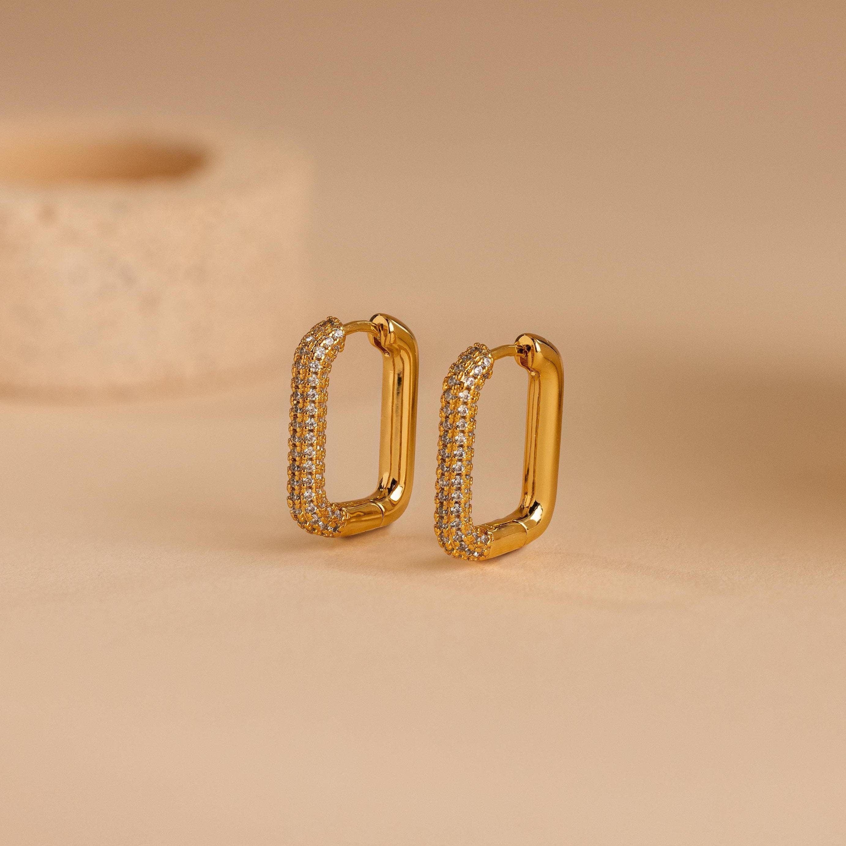 Chunky Pave Link Earrings featuring gold geometric hoops with sparkling pavé stones, displayed upright on a beige surface against a blurred background.