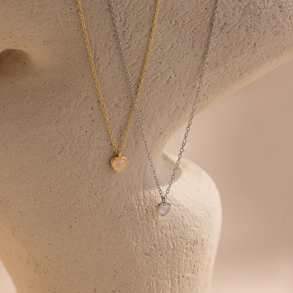 The Tiny Rose Quartz Heart Necklace, shown alongside gold and silver heart-shaped pendant necklaces, is displayed on a textured beige surface.