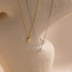 The Tiny Rose Quartz Heart Necklace, shown alongside gold and silver heart-shaped pendant necklaces, is displayed on a textured beige surface.