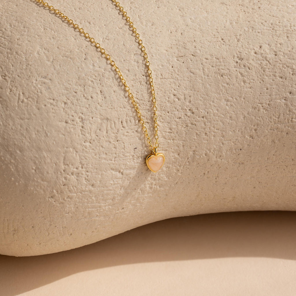 The Tiny Rose Quartz Heart Necklace features a delicate heart-shaped rose quartz pendant set on a gold chain, displayed on a textured beige surface.