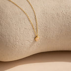 The Tiny Rose Quartz Heart Necklace features a delicate heart-shaped rose quartz pendant set on a gold chain, displayed on a textured beige surface.