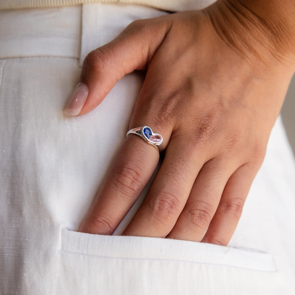 A hand wearing the Duo Birthstone Heart Signet Ring, adorned with blue and pink stones, rests gently on the pocket of white pants.
