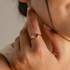 A person wearing the Duo Birthstone Heart Signet Ring, featuring a gold band with a red heart-shaped and a clear round gemstone on their finger.