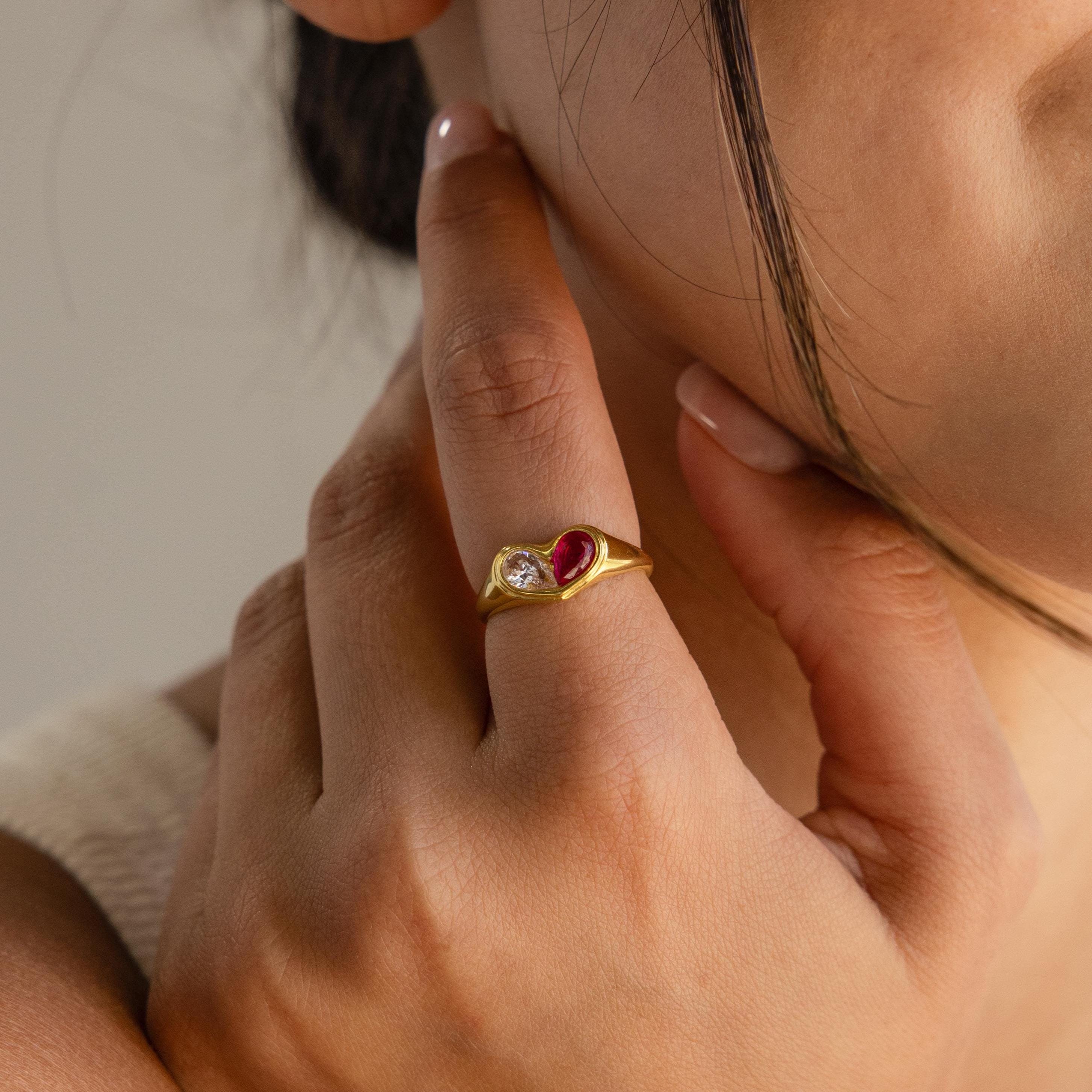 A person wearing the Duo Birthstone Heart Signet Ring, featuring a gold band with a red heart-shaped and a clear round gemstone on their finger.