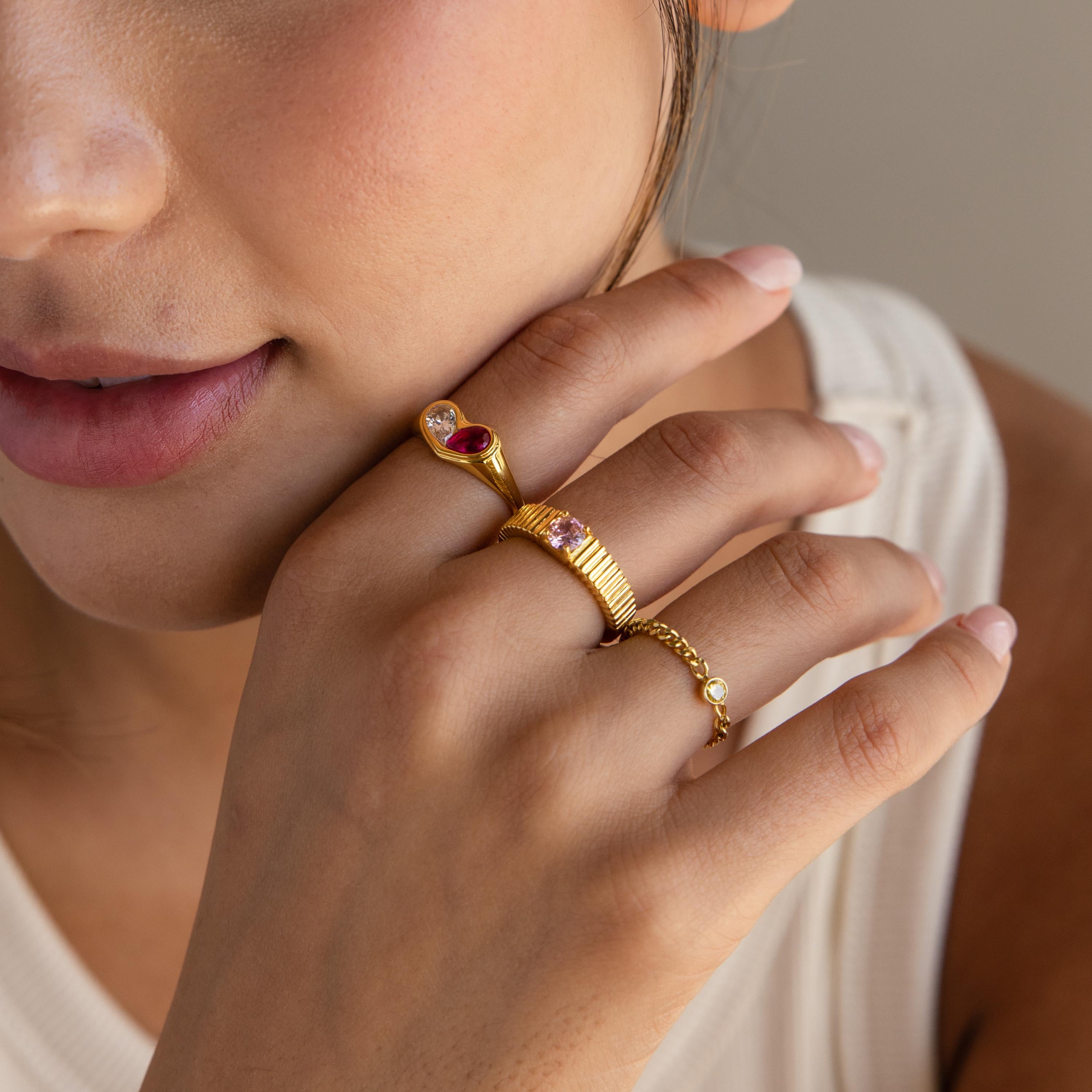 Close-up of a woman wearing three gold rings, including the Duo Birthstone Heart Signet Ring with gemstones, as she gently touches her face.