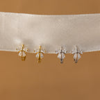 Four pairs of Diamond Ribbon Bow Huggies in 18K Gold, two gold and two silver, displayed on a white ribbon against a tan background.