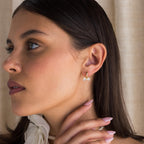 A woman with long dark hair, wearing a white top and the Diamond Bow Huggies, gently touches her neck.