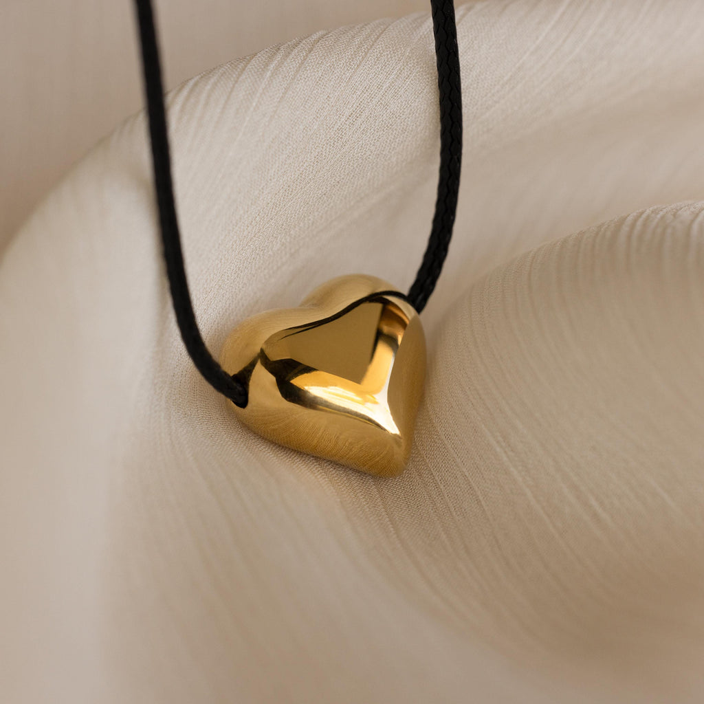 The Chunky Puff Heart Necklace features a gold heart pendant on a black cord, displayed on soft cream fabric—a bold yet elegant statement piece.