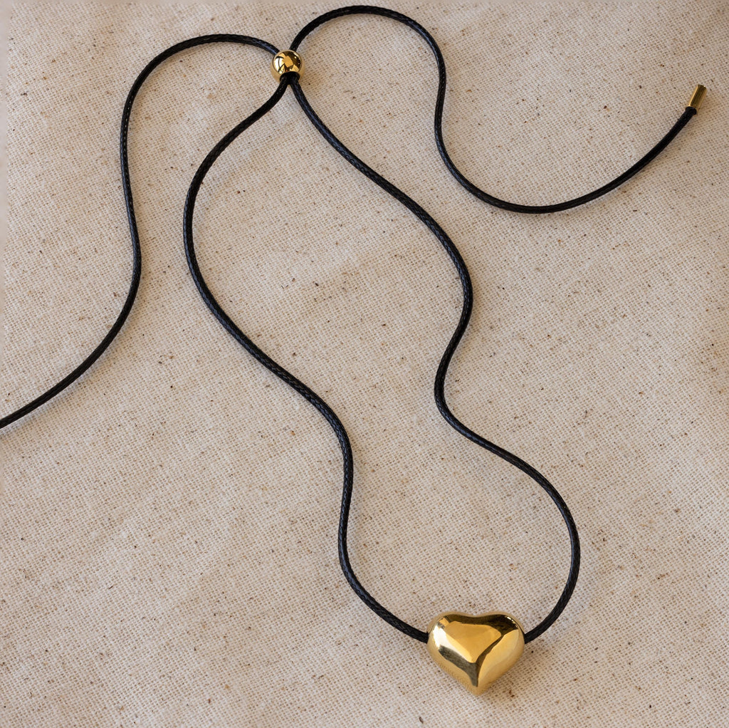 Chunky Puff Heart Necklace with a black cord and shiny gold heart pendant, displayed on beige fabric—perfect for those who love bold heart jewelry.