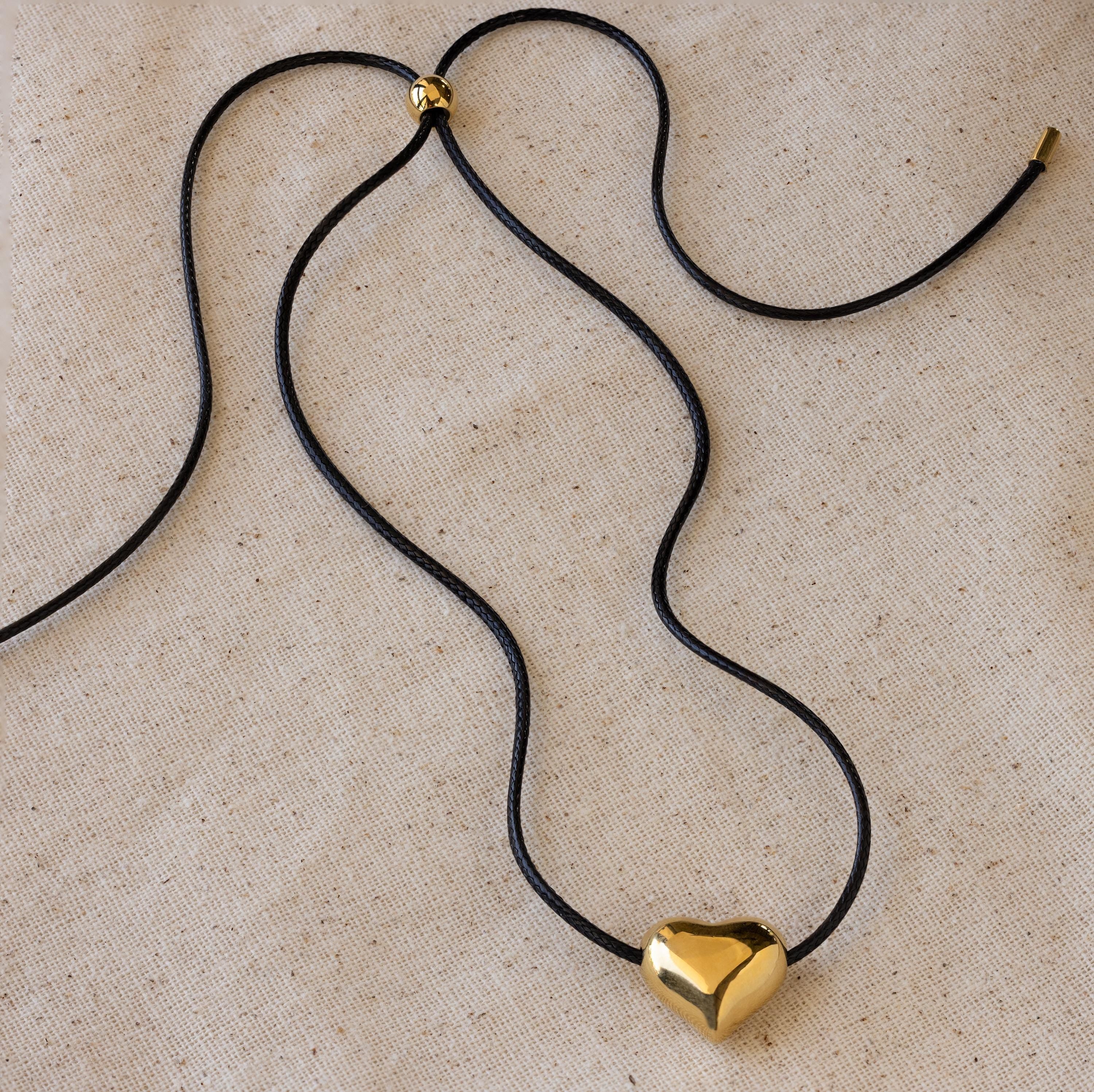 Chunky Puff Heart Necklace with a black cord and shiny gold heart pendant, displayed on beige fabric—perfect for those who love bold heart jewelry.