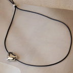 The Chunky Puff Heart Necklace features a black cord and shiny silver heart pendant, displayed on a neutral textured background—a modern twist on the classic heart necklace.