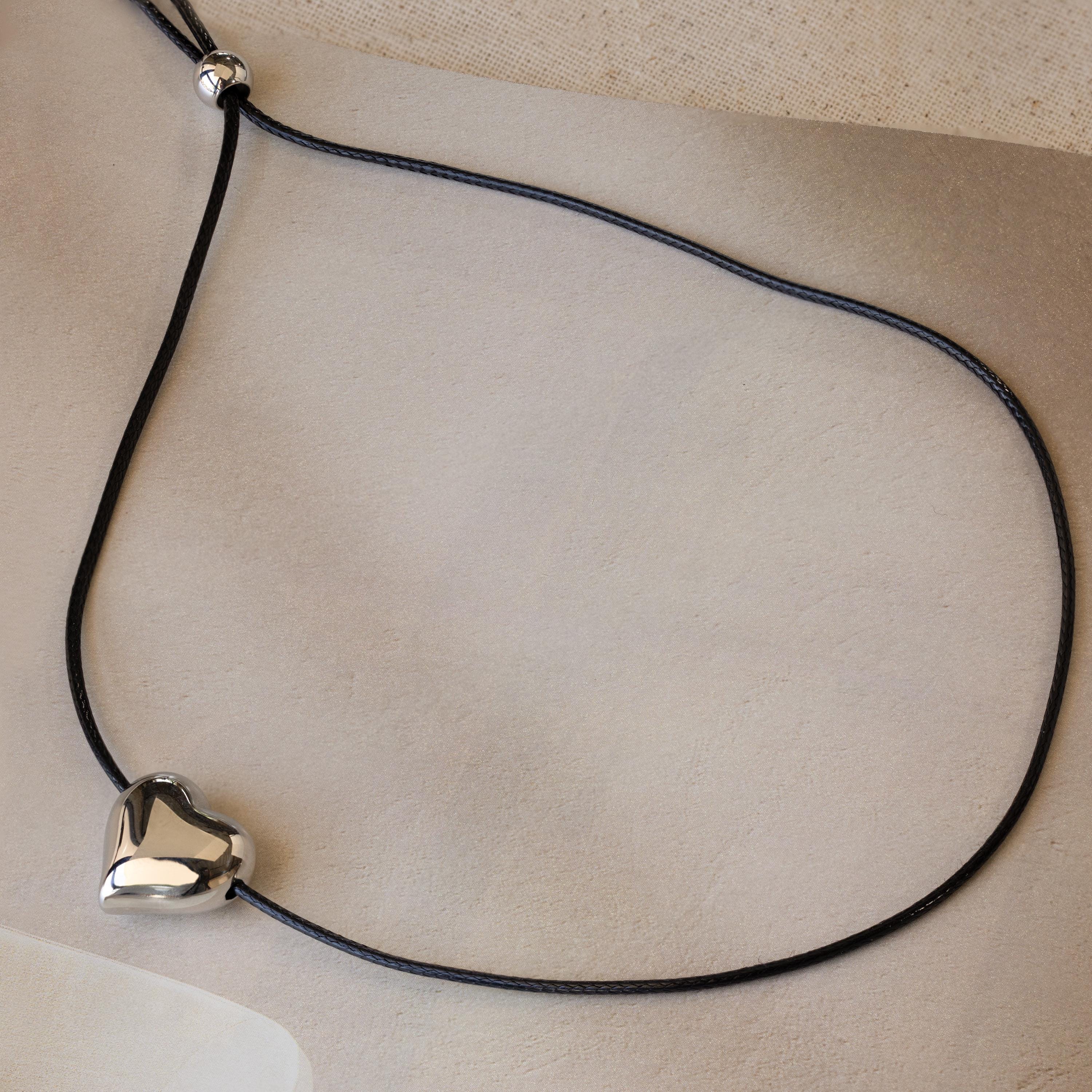 The Chunky Puff Heart Necklace features a black cord and shiny silver heart pendant, displayed on a neutral textured background—a modern twist on the classic heart necklace.