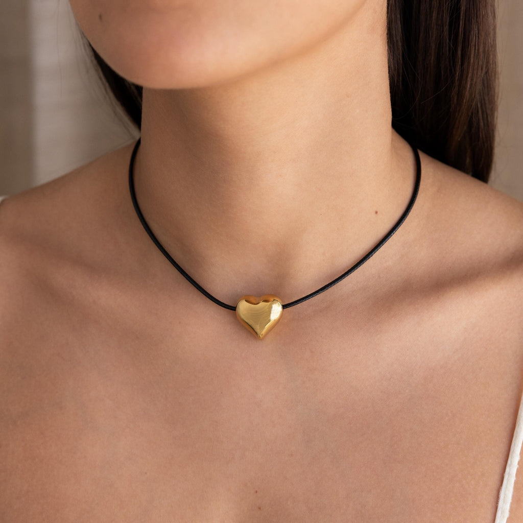 A woman wears the Chunky Puff Heart Necklace, its gold heart pendant on a black cord resting elegantly against her bare skin.