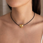 A woman wears the Chunky Puff Heart Necklace, its gold heart pendant on a black cord resting elegantly against her bare skin.