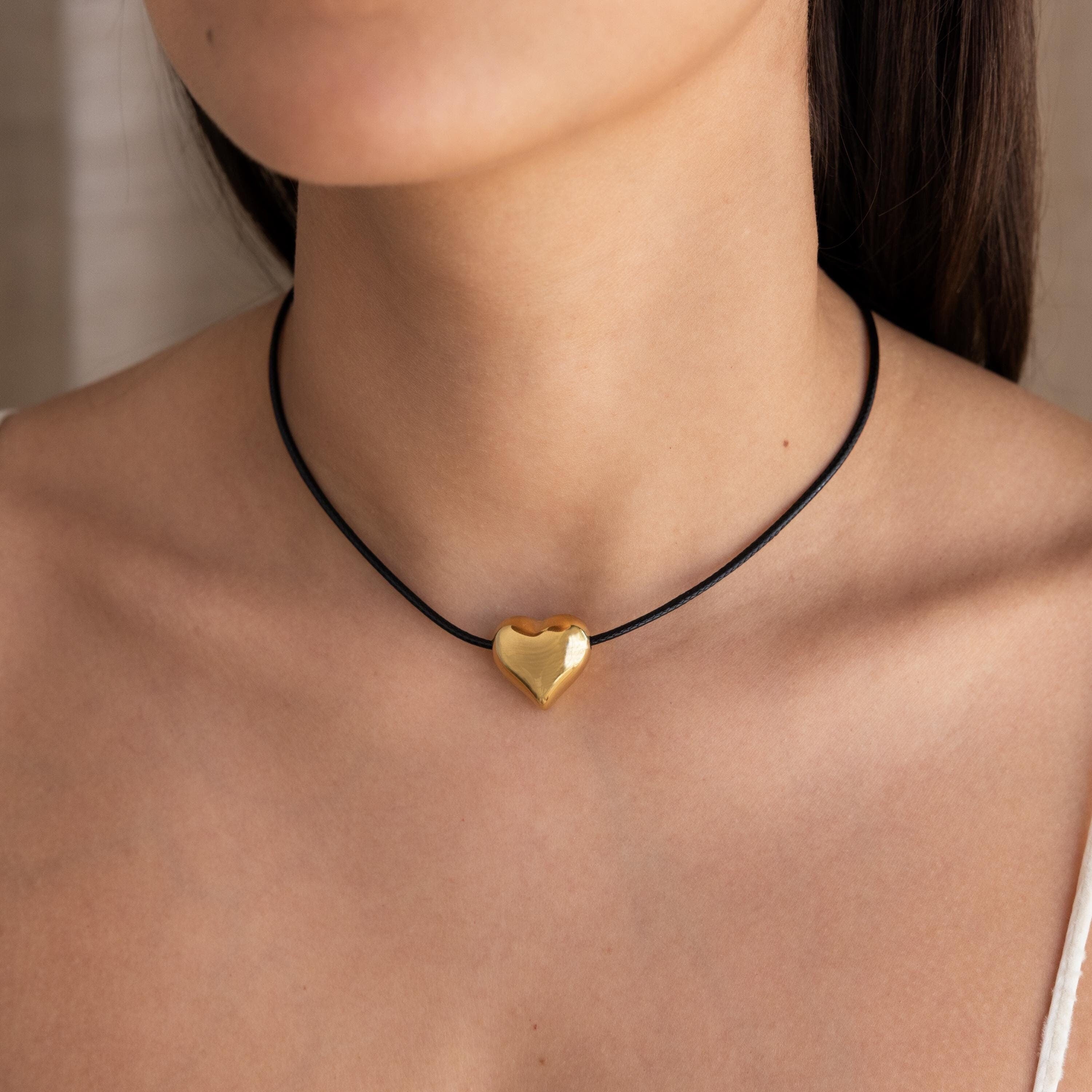 A woman wears the Chunky Puff Heart Necklace, its gold heart pendant on a black cord resting elegantly against her bare skin.