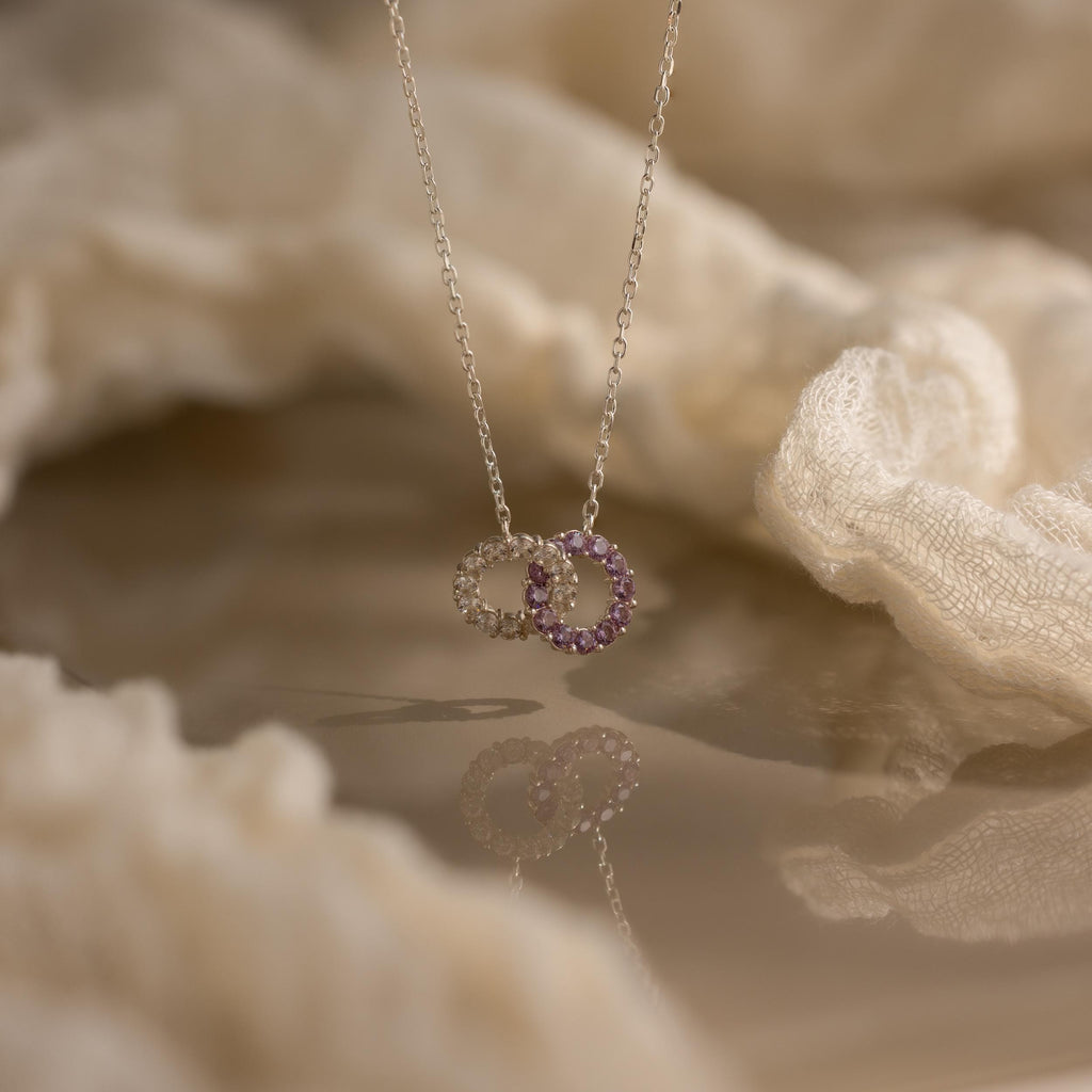 The Interlocking Duo Birthstone Necklace showcases two interlocking circle pendants, one accented with purple stones, displayed on a soft fabric background.