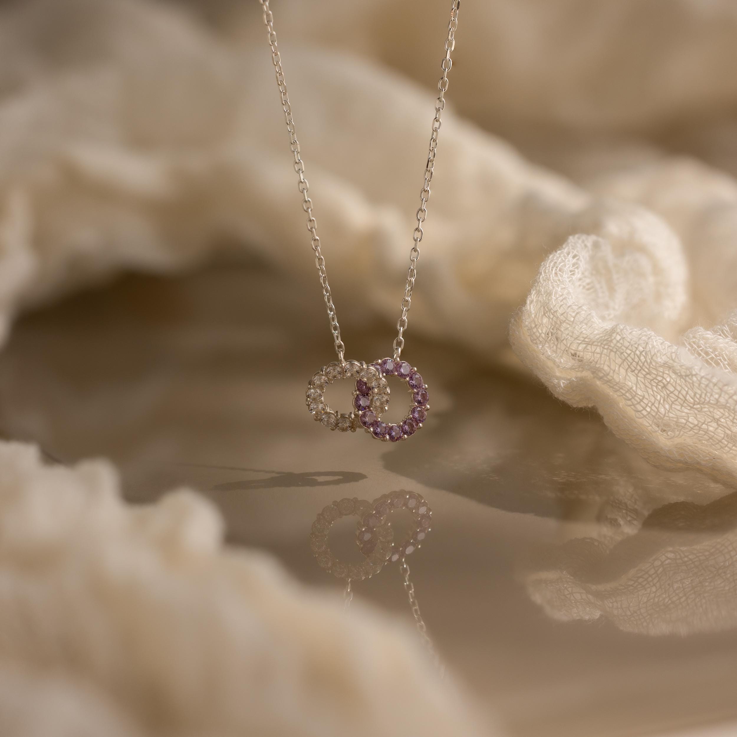 The Interlocking Duo Birthstone Necklace showcases two interlocking circle pendants, one accented with purple stones, displayed on a soft fabric background.