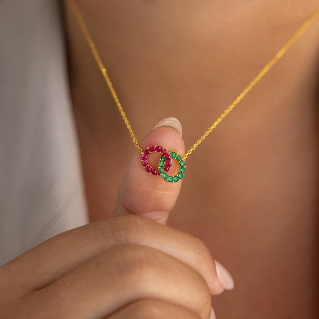 A hand holds the Interlocking Duo Birthstone Necklace, showcasing two interlocking rings adorned with pink and green stones—a beautiful birthstone necklace.