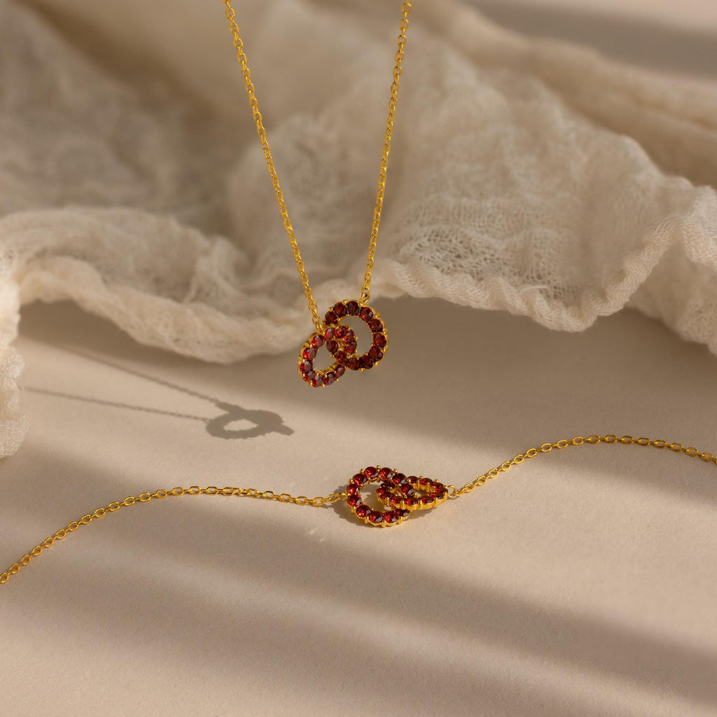The Interlocking Duo Birthstone Necklace features gold circles intertwined with red gemstones, displayed on a soft cream fabric background.