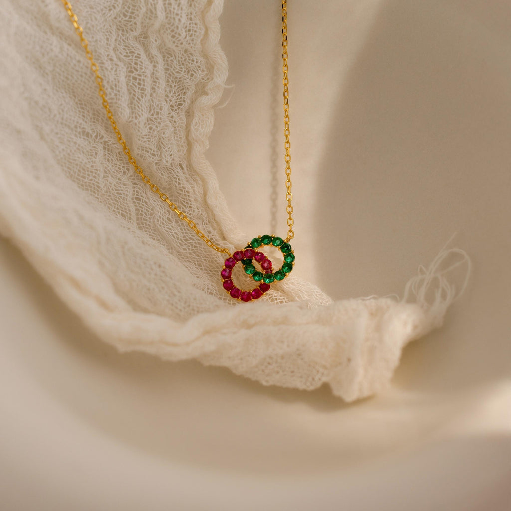 The Interlocking Duo Birthstone Necklace features two gem-studded rings—one green, one pink—beautifully displayed on elegant white lace fabric.