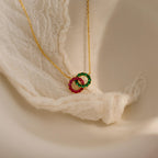 The Interlocking Duo Birthstone Necklace features two gem-studded rings—one green, one pink—beautifully displayed on elegant white lace fabric.