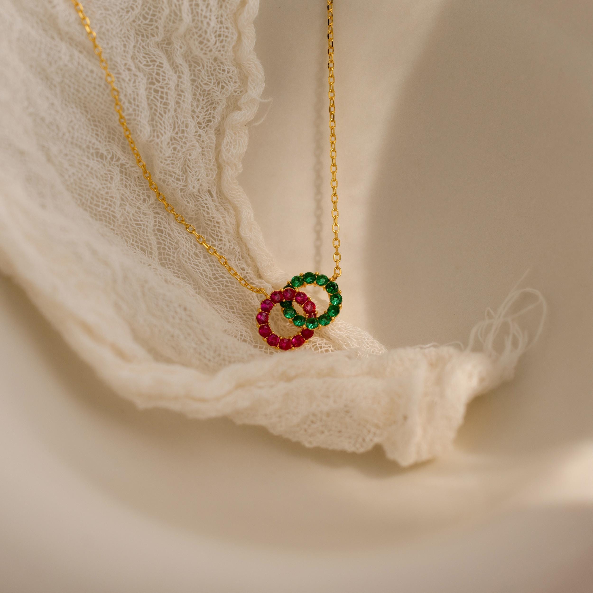 The Interlocking Duo Birthstone Necklace features two gem-studded rings—one green, one pink—beautifully displayed on elegant white lace fabric.