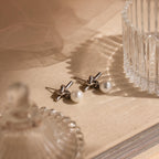 Love Knot Pearl Earrings, silver studs with pearl drops, rest on a beige surface beside clear glass containers and soft fabric—a timeless wedding accessory.