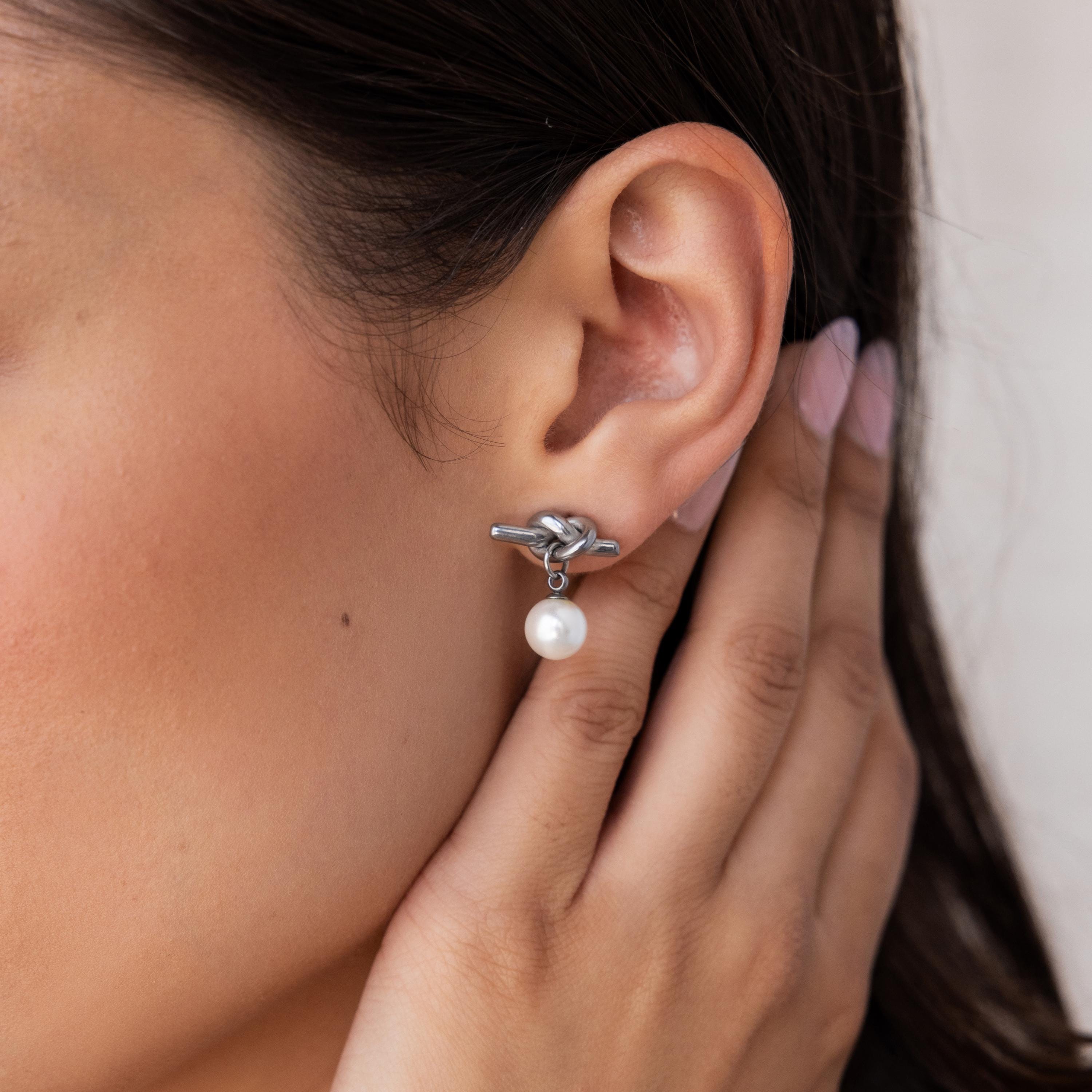 A woman's hand touches her ear, adorned with Love Knot Pearl Earrings featuring a timeless silver knot and an elegant hanging pearl—the perfect accessory for weddings.